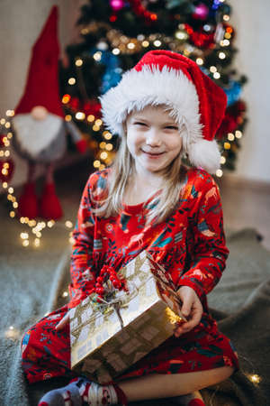 A girl in a hat of santa claus sorts out gifts against the background of a Christmas treeの写真素材