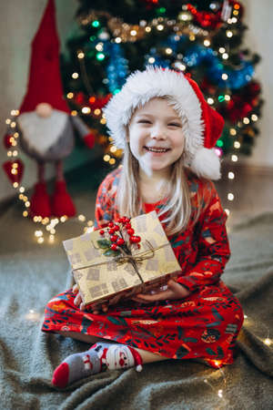 A girl in a hat of santa claus sorts out gifts against the background of a Christmas treeの写真素材