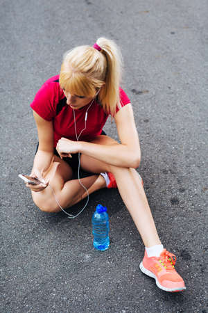 Young pretty runner sitting on the ground and choosing songs to keep her company during the run