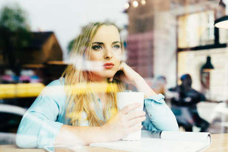 Beautiful young woman drinking coffee at cafe and looking through windowの写真素材