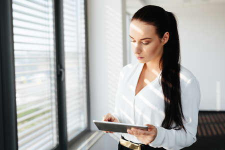 Female manager standing at a window and using her tablet to compare dataの写真素材