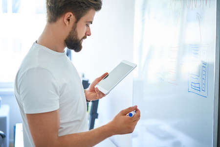 Web designer looking at smartphone while writing notes on whiteboard in modern officeの写真素材