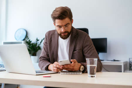 Young businessman checking phone while working with laptop in home officeの写真素材