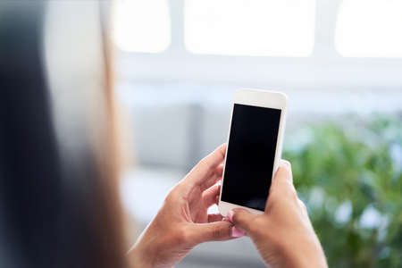 Woman looking at phone while leaning on desk with laptop in home officeの写真素材