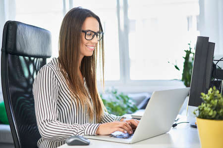 Woman looking at phone while leaning on desk with laptop in home officeの写真素材