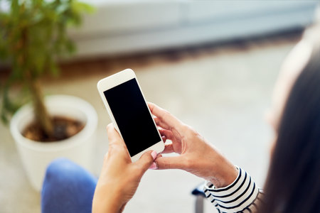 Woman looking at phone while leaning on desk with laptop in home officeの写真素材