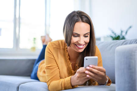 Woman looking at phone while leaning on desk with laptop in home officeの写真素材