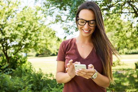 Woman looking at phone while leaning on desk with laptop in home officeの写真素材