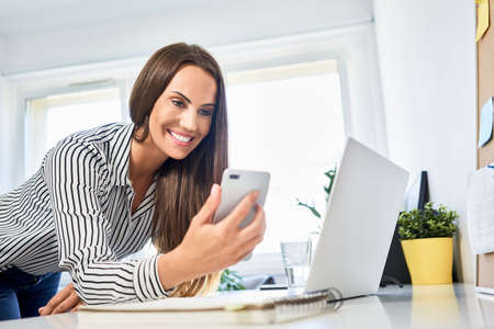 Woman looking at phone while leaning on desk with laptop in home officeの写真素材