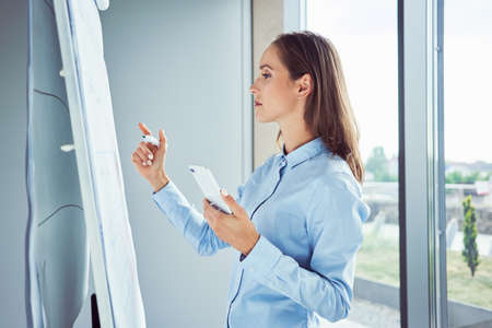 Woman looking at phone while leaning on desk with laptop in home officeの写真素材
