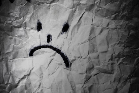 Dissapointed smile. Sad face on crumpled paper in dark background. Depression treatement conceptの写真素材