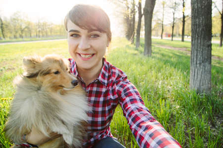 Young woman making selfie with her dog sheltie in the park on the green grass shetland sheepdogの写真素材