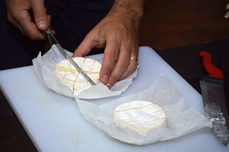 Chef hands cutting wheels of freshly unwrapped camembert cheese, on white cutting board with cheese knife in kitchen.の写真素材