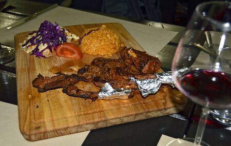 Wooden cutting board with grilled lamb ribs barbecued next to cooked rice and vegetable salad with a nice glass of red wine in the foregroundの写真素材
