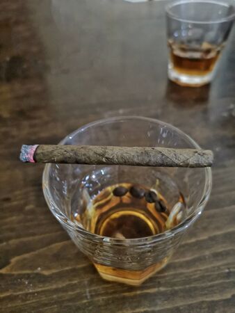 Close up shot of half full glass of whisky on top of wooden bar with lit cigar on top of it, why smoking and alcoholism is bad, sharing scotch whiskey glasses in the pubの写真素材