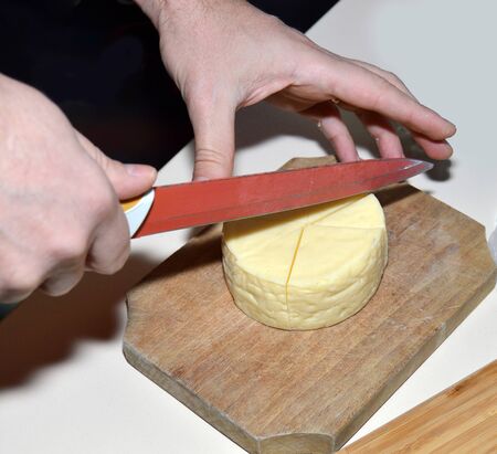 Man hands cutting in half a fresh wheel of tasty organic cheese, slicing cheeses on wooden board for tasting, preparing healthy snackの写真素材