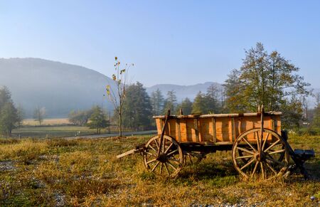 Rural landscape, somewhere in the mountains with warm sunrise light over a field with grass and trees, an old wooden cart with vintage wheels, rustic sceneryの写真素材