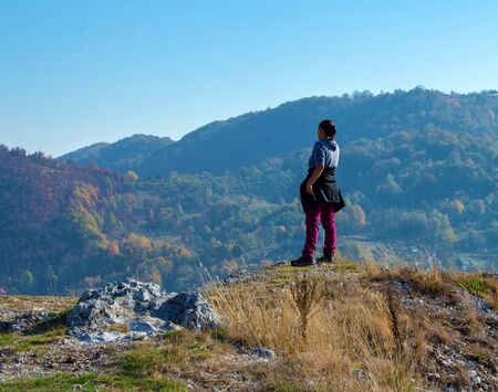 Woman standing on top of a hill, staring into the distance towards the mountains, serene landscape while hiking through the romanian countryside, trekking in natureの写真素材