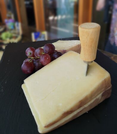 Big block of hard cheese stabbed with cheese cutting instrument or knife, on a wooden platter paired with red grapes ready for tasting and blurry backgroundの写真素材