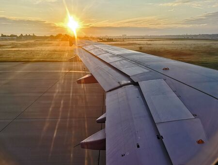 View of plane wing while taking of or landing on airstip / asphalt runaway, view from airplane window with sun reflecting through clouds at dusk or morning, metal plane wingの写真素材