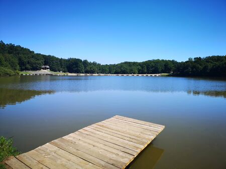 A wooden pontoon or deck over a body of water (lake) surrounded by green trees under a blue sky that's reflected in the watery surface, on a clear summer dayの写真素材