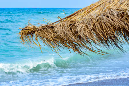 Close-up of straw beach umbrellas providing shade near the wavy water of the Black Sea on sandy shoreline on a sunny clear summer day at Vama Veche, Romaniaの写真素材