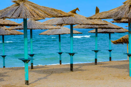 Close-up of straw beach umbrellas providing shade near the wavy water of the Black Sea on sandy shoreline on a sunny clear summer day at Vama Veche, Romaniaの写真素材