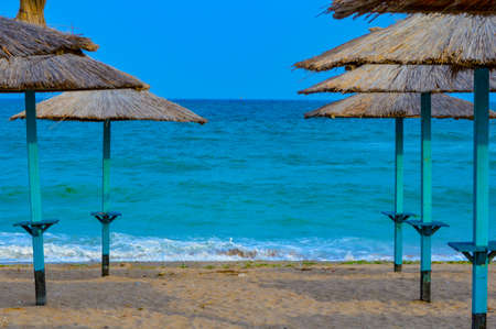 Close-up of straw beach umbrellas providing shade near the wavy water of the Black Sea on sandy shoreline on a sunny clear summer day at Vama Veche, Romaniaの写真素材