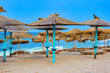 Close-up of straw beach umbrellas providing shade near the wavy water of the Black Sea on sandy shoreline on a sunny clear summer day at Vama Veche, Romaniaの写真素材