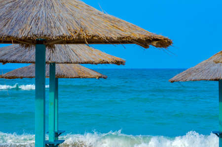 Close-up of straw beach umbrellas providing shade near the wavy water of the Black Sea on sandy shoreline on a sunny clear summer day at Vama Veche, Romaniaの写真素材