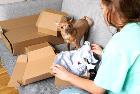 Unpacking cardboard boxes concept. Pet owner and small dog sitting on sofa and unboxing cardboard boxes.の写真素材