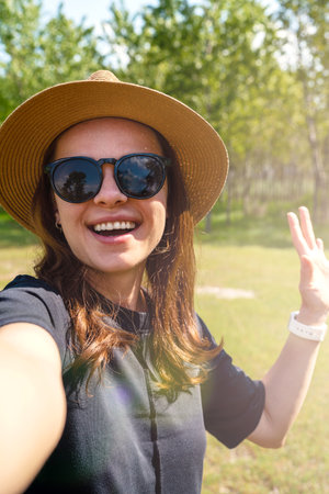 Woman wearing sunglasses and straw hat waving hand and taking a selfie while walking outdoors.の写真素材