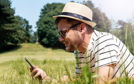 Stylish man lying down on green lawn in the city park and chatting online using his phone.
