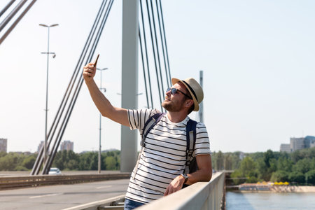 A male tourist with a backpack in sunglasses and a straw hat stands on a bridge over the river and takes a selfie using his phone.の写真素材