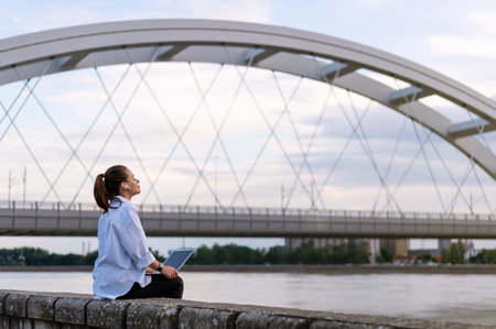Urban woman with her laptop sitting on the waterfront on background of river bridge, end of the working day.の写真素材