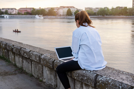 Woman working remotely on her laptop outside the office sitting on waterfront opposite the river European city.の写真素材