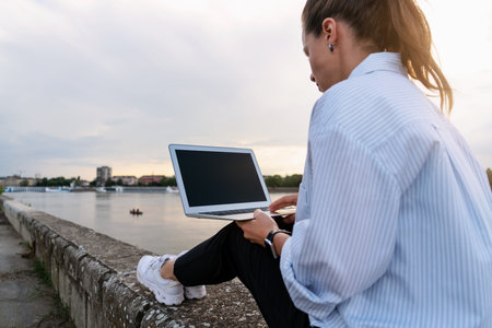 Urban woman sitting on the city waterfront and using her laptop, meeting the end of the work day out of office.の写真素材