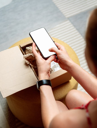 Female person customer of internet store unpacking online order and taking a photo of goods using smartphone with blank screen.の写真素材