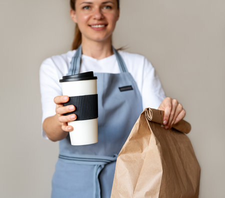 Compostable coffee cup and paper bag with takeaway meal in hands of pretty waitress. Eco-friendly package for takeout food.の写真素材