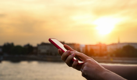 Female person touching screen of her phone with her finger in front of background of city during sunset.の写真素材