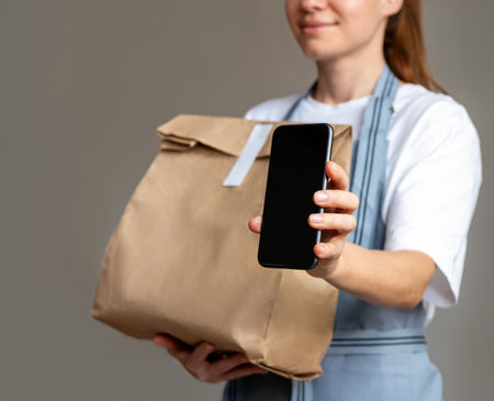 Waitress in apron holding paper bag with takeout food and showing screen of smartphone. Mobile payment with qr-code, food online ordering.の写真素材