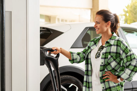 Woman at an EV charging station in the city, charging her electric car. Clean energy, electrification, and environmental awareness.の写真素材