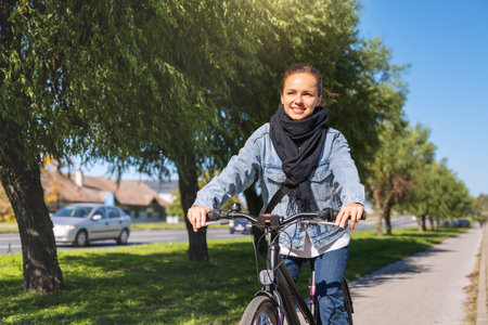 Urban woman in a denim jacket and black scarf riding a bicycle along a city bike path, with green trees in the background.の写真素材