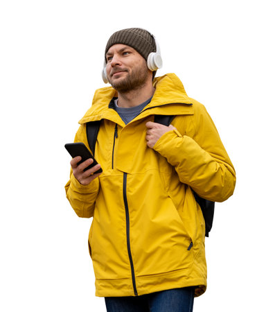 Caucasian man in a yellow winter jacket and hat, standing with a mobile phone, wireless headphones,...