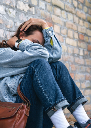 Stressed woman curling up hiding her face with her hands next to brick wall outdoors on city street.の写真素材