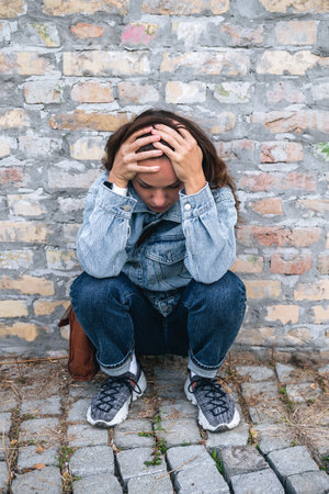 Female in emotional pain sitting and holding head in hands against brick wall on streetの写真素材