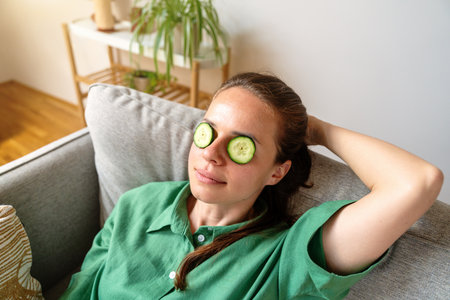 Peaceful woman enjoying home spa moment with cucumber eye mask on sofaの写真素材