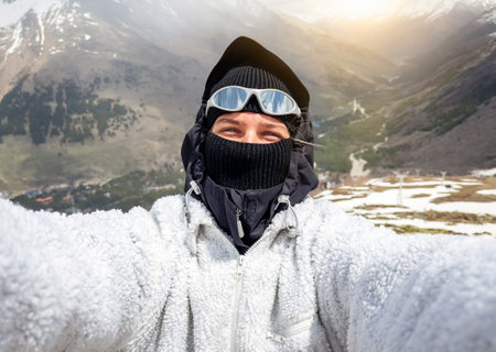 Female hiker in warm clothing and mask taking selfie with mountains in background
