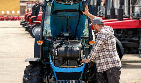 Agricultural worker examining tractor with open hood in outdoor farm machinery lot