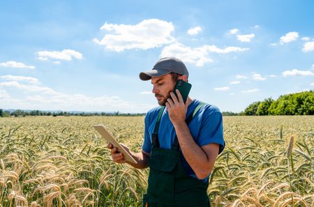 Multitasking male farmer talking on phone and using tablet while standing in cereak field in...